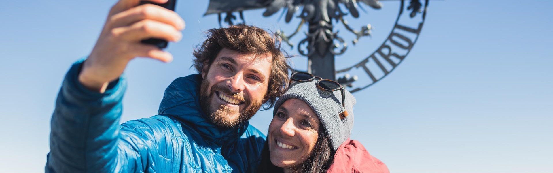 Paar macht ein Selfie mit einem Gipfelkreuz im Hintergrund